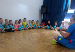 dzieci siedzą z piłkami pod ścianą i przed instruktorem
