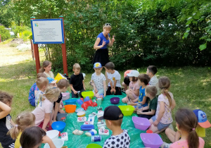dzieci siedzą wokół folii rozłożonej na trawie w ogrodzie a pani Aneta podaje instrukcje wykonania pracy