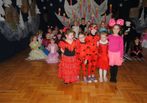 dzieci siedzą z tyłu na podłodze a grupa dziewczynek w czerwonych kostiumach, w czarne kropki prezentuje się przed nimi