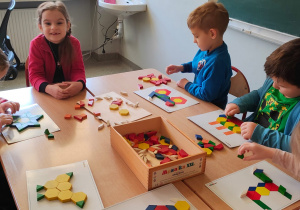 dzieci siedzą przy stoliku i układają drewniane klocki o różnych wzorach i kolorach na kartkach z gotowym wzorem