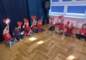 przedszkolaki siedzą w kole i trzymają papierowe serca nad głową