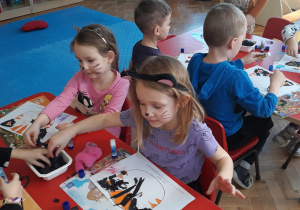 przedszkolaki wykonują prace plastyczne przy stolikach - wyklejają kontur twarzy kota czarną i pomarańczową bibułą