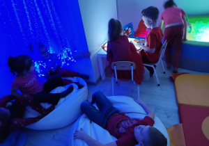 przedszkolaki w sali doświadczania świata przy podświetlanym stoliku a obok dzieci relaksują się pod świetlistą chmurką
