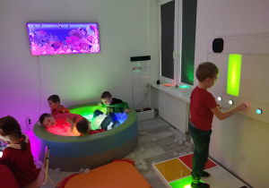 przedszkolaki w sali doświadczania świata w podświetlanym basenie z kulkami a obok chłopiec doświadcza dźwięków wydobywanych nogami