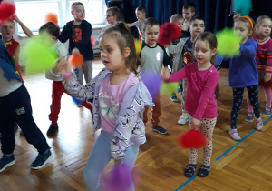 przedszkolaki tańczą ustawione w szeregach z kolorowymi pomponami, machając jednym z nich w górze