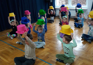 dzieci siedzą skrzyżnie na podłodze trzymając kolorowe kapelusze nad głową