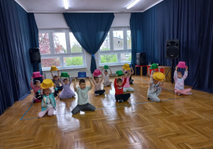 przedszkolaki siedzą skrzyżnie na podłodze z uniesionymi rękami nad głowami z kolorowymi kapeluszami