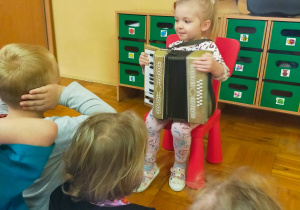 dziewczynka siedzi na krzesełku przed grupą przedszkolaków i trzyma akordeon na kolanach