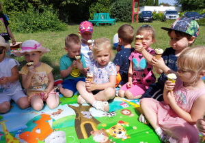 przedszkolaki jedzą lody siedząc na macie w ogrodzie przedszkolnym