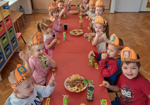 przedszkolaki podczas słodkiego poczęstunku w pomarańczowych czapkach na głowie z napisem "Dzien Przedszkolaka"