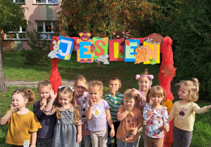 przedszkolaki z grupy VI pozują do zdjęcia pod bramką z napisem "jesień" ustawioną w ogrodzie przedszkolnym, na tle budynku przedszkola i jarzębiny