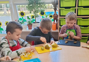 chłopcy podczas krojenia sałatki owocowej siedzą przy stoliku i kroją owoce na deskach