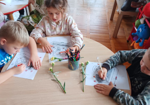 przedszkolaki kolorują a nad obrazkiem leżą goździki związane wstążką na kokardkę