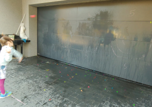 dziewczynka stoi przed garażem i rzuca w jego drzwi balony wypełnione wodą