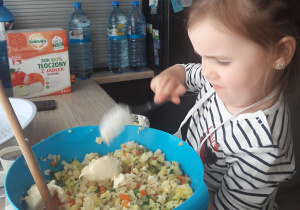 dziewczynka wkłada łyżką majonez do niebieskiej miski z pokrojoną sałatką