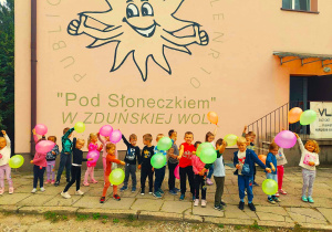 przedszkolaki z balonami przed budynkiem przedszkola z narysowanym logo placówki słoneczkiem