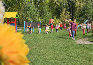 przedszkolaki idą w korowodzie za nauczycielką po ogrodzie przedszkolnym