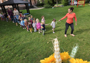 przedszkolaki idą w korowodzie za nauczycielką po ogrodzie przedszkolnym