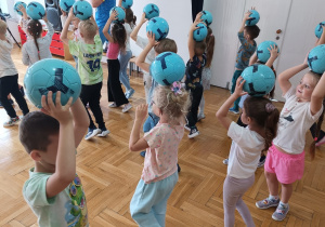 dzieci trzymają piłki nad głowami i maszerują po sali