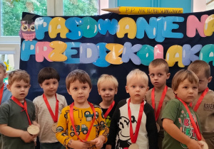 zdjęcie grupowe chłopcówz zawieszonymi na szyi medalami przed tablicą z napisem pasowanie na przedszkolaka