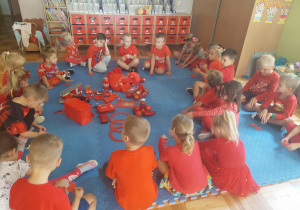 dzieci siedzą w kole zgromadzone wokół czerwonych przedmiotów zgromadzonych na macie
