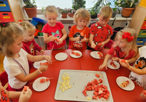 dzieci przy stoliku nakłuwają kawałki arbuza i sra żółtego robiąc szaszłyki