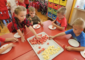 dzieci przy stoliku nakłuwają kawałki arbuza i sra żółtego robiąc szaszłyki