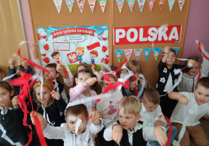 przedszkolaki machają biało czerwonymi wstążkami przed tablicą z napise m Polska, ilustracją symboli narodowych oraz wykonanymi przez dzieci proporczykami z napisem 11 listopada