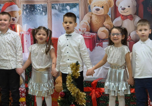 dzieci ustawione wokół choinki i prezentów śpiewają trzymając się za ręce