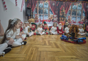 dzieci przebrane za śnieżynki i skrzaty siedzą w półkolu wokól choinki i śpiewają