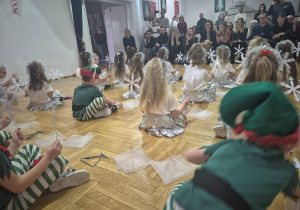 przedszkolaki wykonują układ taneczno-rytmiczny ze śnieżynkami siedząc przed rodzicami