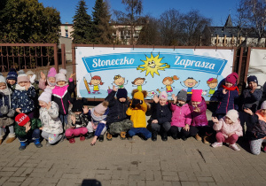 przedszkolaki siedzą pod kolorowym banerem z napisem "Słoneczko zaprasza" zawieszonym na ogrodzeniu placówki